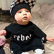 Ryder is registered to the contest to win money with this photo: baby, child, infant, black_clothing, checkered_pattern, cap, socks, bed, blanket, pacifier, cat, indoor, curious_expression, sitting, person, face, headwear, cute, home, soft_lighting