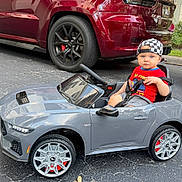 Ryder is registered to the contest to win money with this photo: toddler, child, toy_car, cap, checkered, red_shirt, gray_car, parking_lot, driveway, suv, red_suv, wheel, steering_wheel, outdoor, asphalt, vehicle, young_child, playing, casual, daytime