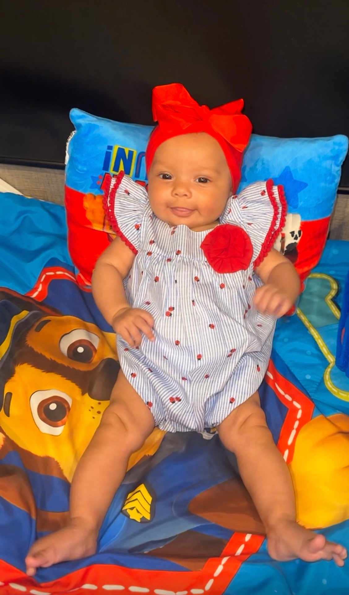 Kinlee joined the competition — help win amazing prizes! baby, child, infant, smiling, headband, red_bow, striped_outfit, ladybug_pattern, pillow, blanket, paw_patrol, colorful, cute, sitting, indoors, portrait, happy, person, young_child, playful