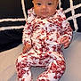 Kinlee is registered to the contest to win money with this photo: baby, infant, onesie, floral_pattern, sitting, bed, blanket, face, portrait, child, cute, indoors, person, clothing, pattern, background, soft_toys, young_child, head, expression