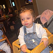 Kyrell is registered to the contest to win money with this photo: toddler, child, high_chair, denim_overalls, white_shirt, indoor, curly_hair, red_eye, wooden_furniture, carpet, person, seated, baby, clothing, face, hand, footwear, family, candid, room