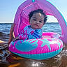 toddler, child, inflatable_float, water, lake, sunny, curly_hair, pink, blue, swimwear, summer, outdoor, water_safety, person, floating, hand, reflection, vacation, relaxation, nature