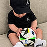 baby, child, infant, soccer_ball, football, sport, sportswear, baseball_cap, black_clothing, jordan, socks, white_socks, sitting, sofa, indoor, portrait, holding_ball, looking_down, textile_background, cute