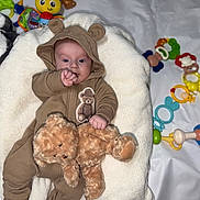 Aaron participe au concours pour gagner de l'argent avec cette photo : baby, infant, onesie, brown_clothing, teddy_bear, plush_toy, cushion, white_background, colorful_toys, pacifier, soft_toy, child, cute, indoors, smiling, face, hand, comfort, playtime, lying_down