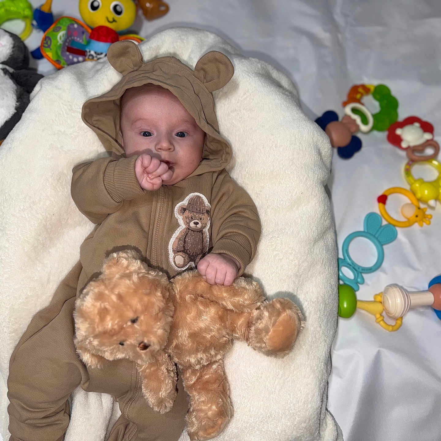 Aaron participe au concours pour gagner de l'argent avec cette photo : baby, infant, onesie, brown_clothing, teddy_bear, plush_toy, cushion, white_background, colorful_toys, pacifier, soft_toy, child, cute, indoors, smiling, face, hand, comfort, playtime, lying_down
