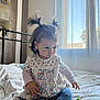 toddler, child, bed, pigtails, toys, window, curtain, natural_light, indoor, clothing, socks, blue_leggings, floral_top, person, cute, young_child, playing, home, soft_light, focused