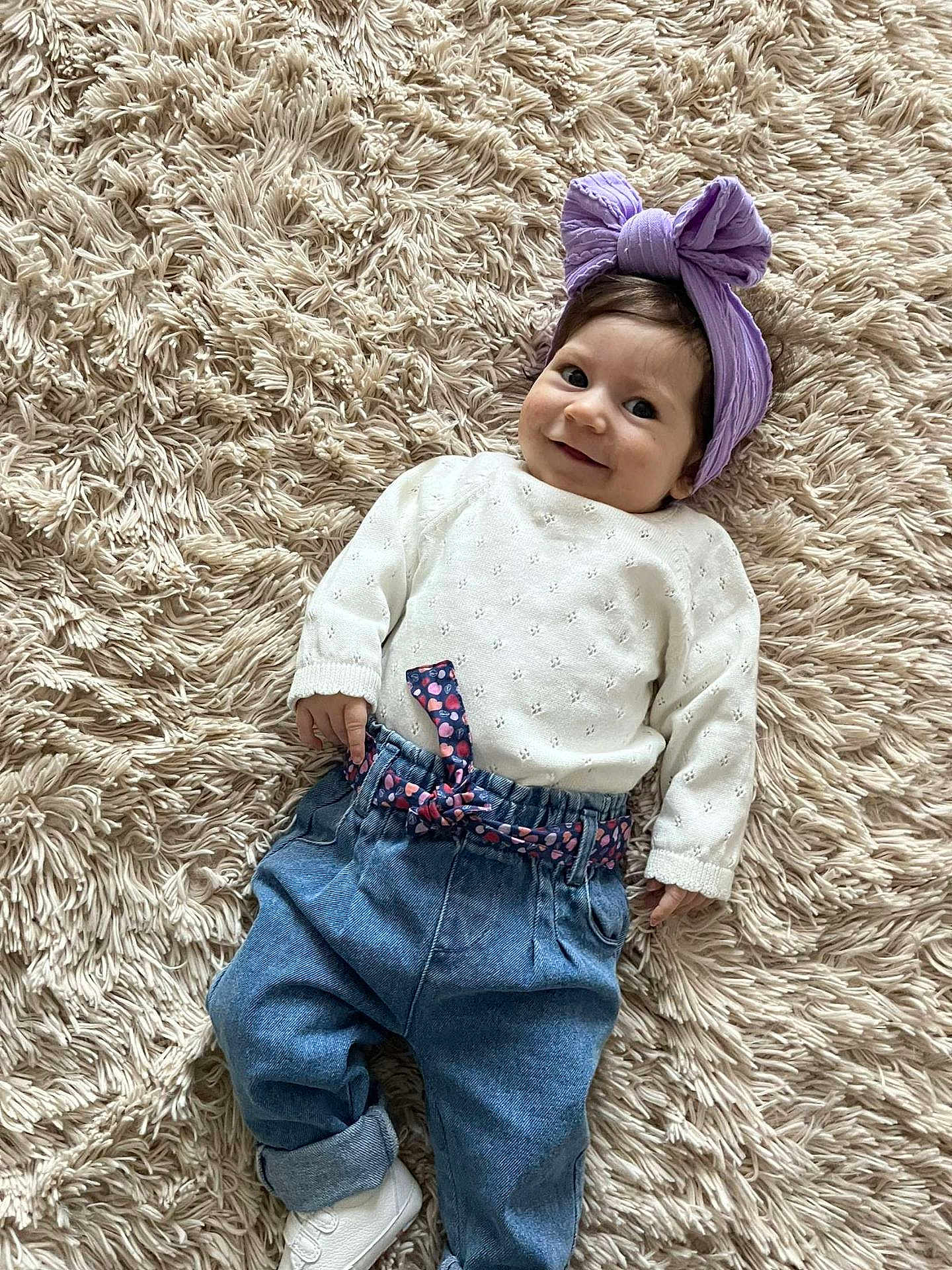Dunja a rejoint le concours — aidez-le/la à gagner de superbes lots ! baby, child, smiling, headband, purple_bow, white_sweater, blue_jeans, belt, carpet, fluffy, indoor, cute, infant, portrait, person, footwear, white_shoes, happy, lying_down, playful