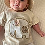 baby, infant, hat, elephant, shirt, clothing, white_pants, blanket, lying_down, cute, child, face, portrait, indoors, soft_light, fabric, texture, smiling, person, small_hand