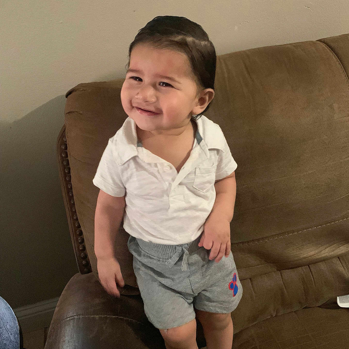 Napoleon is registered to the contest to win money with this photo: brown_couch, casual_clothing, child, couch, cute, face, furniture, grey_shorts, hair, happy, home, indoors, living_room, person, smiling, socks, standing, toddler, white_shirt, young_child