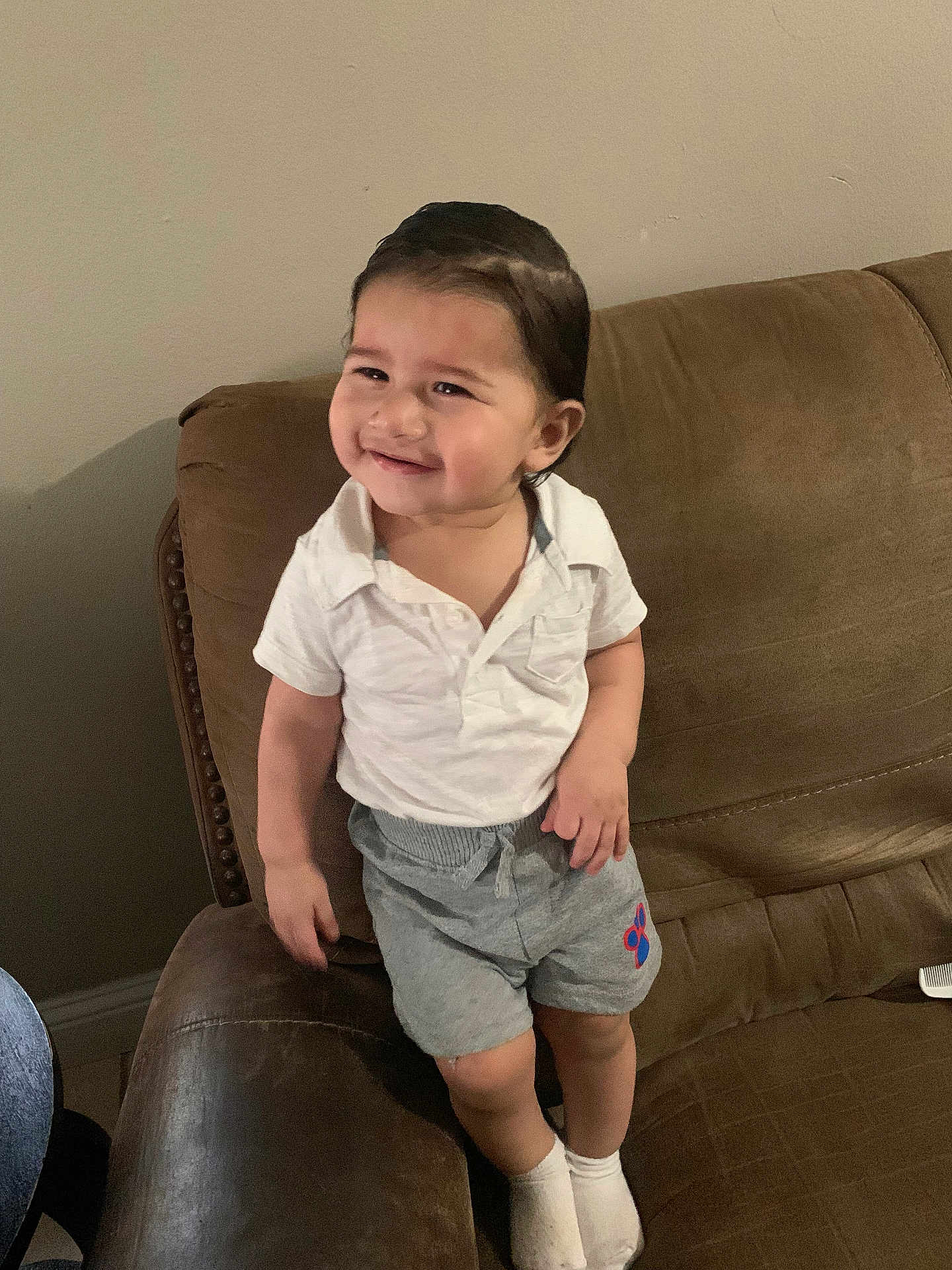 Napoleon is registered to the contest to win money with this photo: brown_couch, casual_clothing, child, couch, cute, face, furniture, grey_shorts, hair, happy, home, indoors, living_room, person, smiling, socks, standing, toddler, white_shirt, young_child