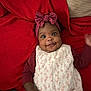 baby, child, infant, smile, headband, bow, red_blanket, floral_dress, happy, cute, portrait, face, eyes, hand, lying_down, indoors, apparel, person, skin, one_person