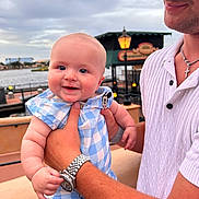 Weston is registered to the contest to win money with this photo: baby, man, smile, holding, blue_checkered_outfit, mickey_mouse_patch, waterfront, lamp_post, cloudy_sky, watch, white_shirt, bracelet, outdoor, hand, portrait, happy, person, skin, face, daylight