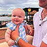baby, man, smile, holding, blue_checkered_outfit, mickey_mouse_patch, waterfront, lamp_post, cloudy_sky, watch, white_shirt, bracelet, outdoor, hand, portrait, happy, person, skin, face, daylight