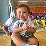Nathan is registered to the contest to win money with this photo: toddler, child, smiling, indoor, table, clapping, person, adult, white_shirt, yellow_chairs, background, happy, shorts, socks, wooden_table, people, casual_clothing, daylight, restaurant, play_area