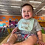 Nathan joined the competition — help win amazing prizes! baby, child, smiling, indoor, bib, plaid_shorts, wooden_table, colorful_chairs, play_area, dinosaur_mural, clouds, happy, cute, person, short_sleeves, feet, hands, infant, expression, casual_clothing