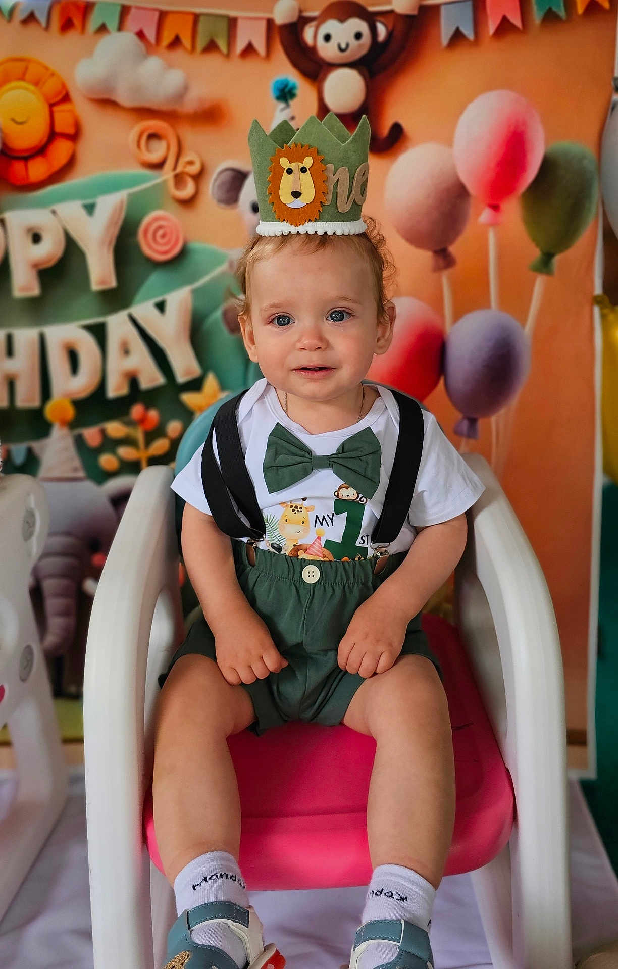 Constatin is registered to the contest to win money with this photo: child, toddler, birthday, party, balloons, crown, green_clothing, suspenders, bow_tie, chair, pink_chair, smiling, celebration, decorations, monkey, background, first_birthday, cute, indoor, happy