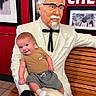 baby, child, infant, smiling, sitting, statue, colonel_sanders, restaurant, bench, wood, red_wall, white_suit, bow_tie, glasses, framed_pictures, indoor, portrait, casual_clothing, sneakers, funny_pose