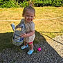 toddler, child, outdoor, grass, gravel, water_bottle, toy, pink, blue_shoes, smiling, person, shorts, shirt, sunlight, shadow, nature, hedge, playing, happy, summer