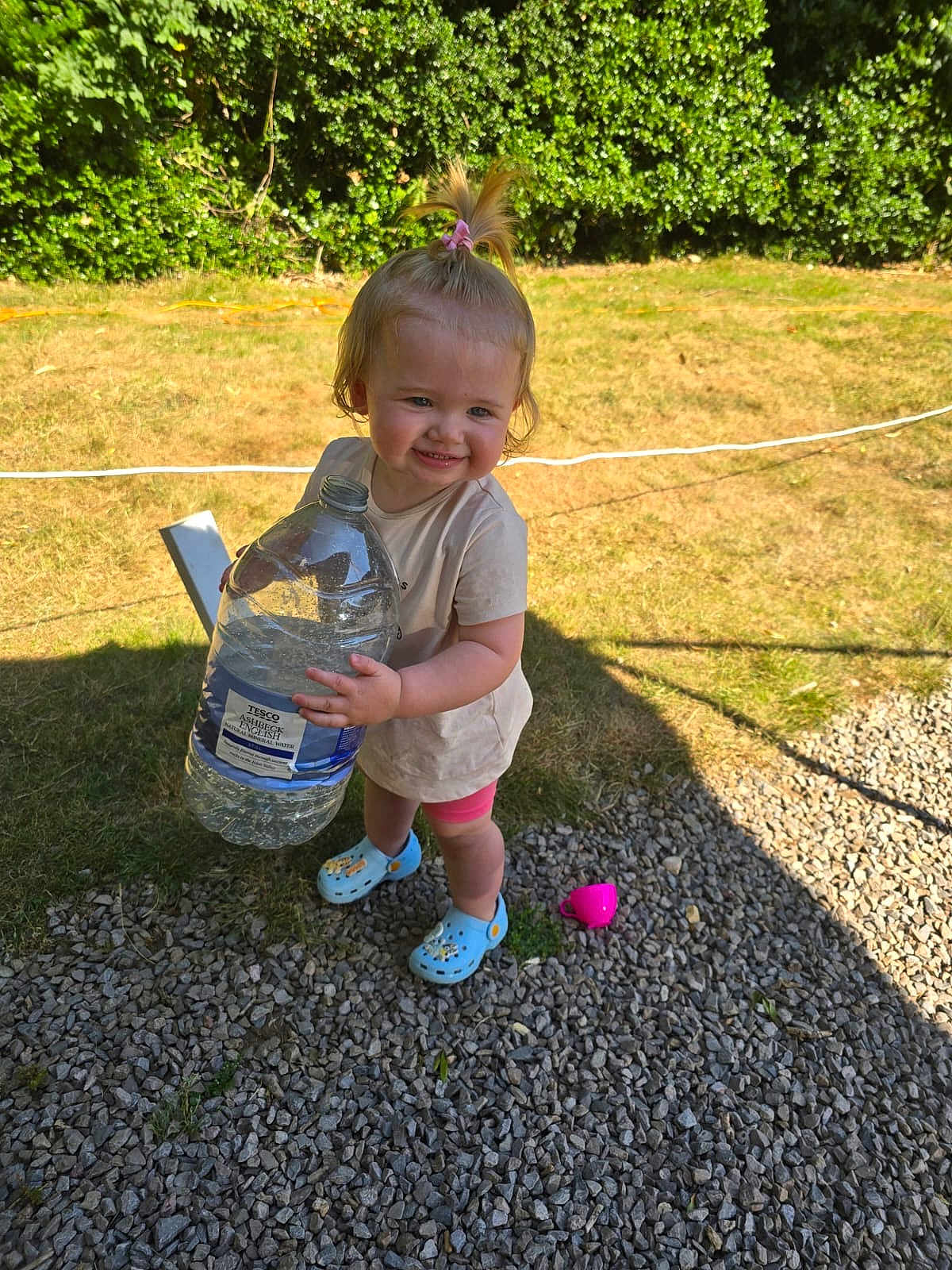 Annabelle is registered to the contest to win money with this photo: toddler, child, outdoor, grass, gravel, water_bottle, toy, pink, blue_shoes, smiling, person, shorts, shirt, sunlight, shadow, nature, hedge, playing, happy, summer