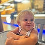 toddler, child, blonde_hair, smiling, indoor, table, chair, face, portrait, person, happy, blue_eyes, casual_clothing, arm, skin, cute, baby, young_child, seated, background_blur
