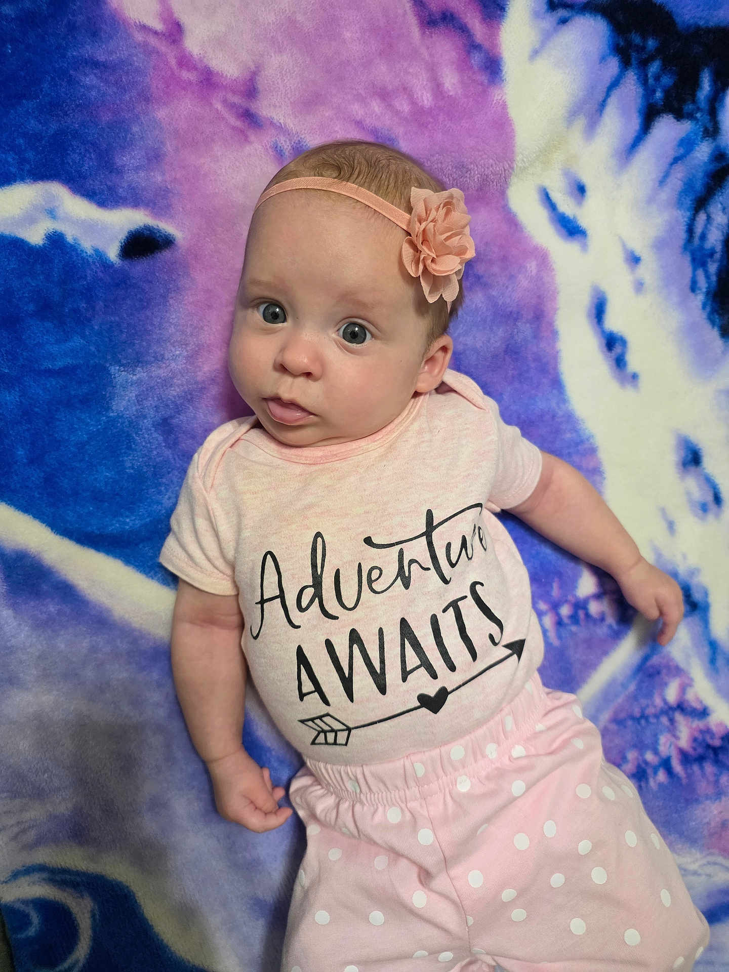 Emily joined the competition — help win amazing prizes! baby, infant, wide_eyes, headband, flower_headband, pink_onesie, text_on_shirt, polka_dot_pants, blanket, purple_background, blue_background, lying_down, portrait, cute, small_hand, skin, staring, soft_texture, nursery, indoors