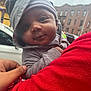baby, child, hoodie, beanie, person, urban, outdoor, car, building, red_clothing, hand, face, portrait, cute, infant, casual_clothing, street, holding, expression, gray_clothing