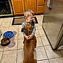 child, dog, hug, kitchen, tile_floor, brown_dog, refrigerator, cabinet, food_bowl, child_hugging_dog, pet, indoor, affection, cute, boy, animal, floor_mat, feeding, love, home