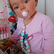 Romane participe au concours pour gagner de l'argent avec cette photo : toddler, child, pacifier, pink_clothing, patterned_pants, toy, blanket, indoor, cute, portrait, face, hand, hair_tie, sitting, young_child, baby_accessory, colorful, playful, home, curious