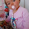 toddler, child, pacifier, pink_clothing, patterned_pants, toy, blanket, indoor, cute, portrait, face, hand, hair_tie, sitting, young_child, baby_accessory, colorful, playful, home, curious