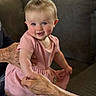 baby, child, toddler, smiling, portrait, indoor, living_room, couch, pink_dress, hair_bow, blue_eyes, sitting, standing, adult_hands, family, home, soft_lighting, blanket, furniture, happy