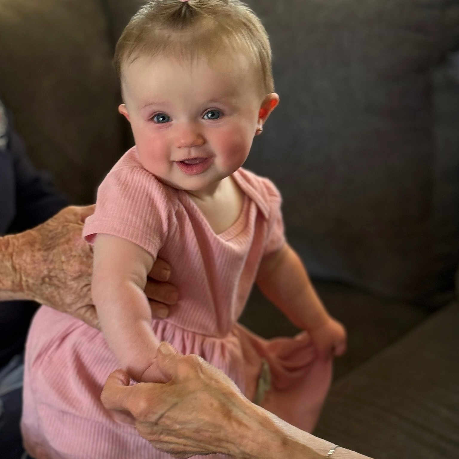 Raija joined the competition — help win amazing prizes! baby, child, toddler, smiling, portrait, indoor, living_room, couch, pink_dress, hair_bow, blue_eyes, sitting, standing, adult_hands, family, home, soft_lighting, blanket, furniture, happy