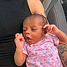 baby, infant, child, person, tattoo, car_seat, seatbelt, black_clothing, pink_onesie, floral_pattern, hand, face, skin, arm, denim, casual, portrait, indoors, relaxed, expression