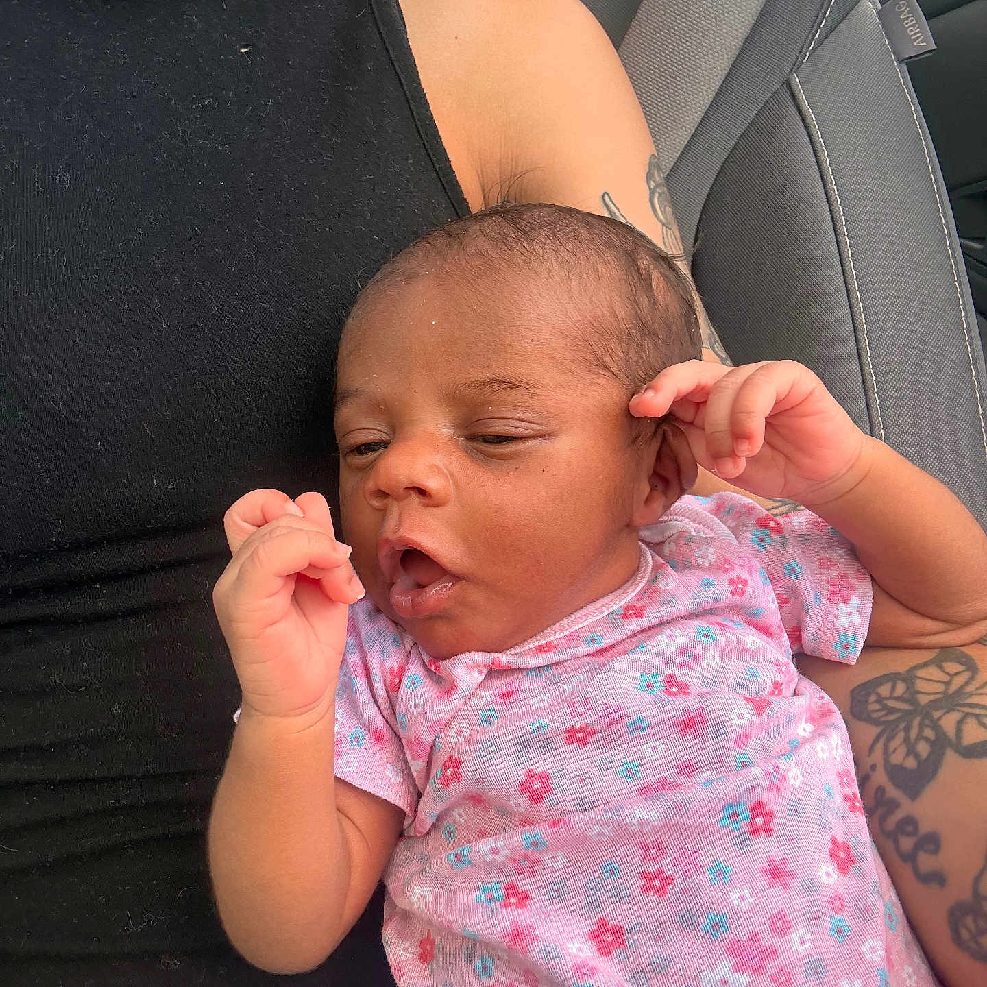 Anastasia is registered to the contest to win money with this photo: baby, infant, child, person, tattoo, car_seat, seatbelt, black_clothing, pink_onesie, floral_pattern, hand, face, skin, arm, denim, casual, portrait, indoors, relaxed, expression
