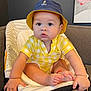 baby, child, hat, bucket_hat, yellow_clothing, baby_seat, indoor, cute, chubby_cheeks, hands, feet, polo_logo, plaid_pattern, seated, portrait, infant, couch, painting, home, curious
