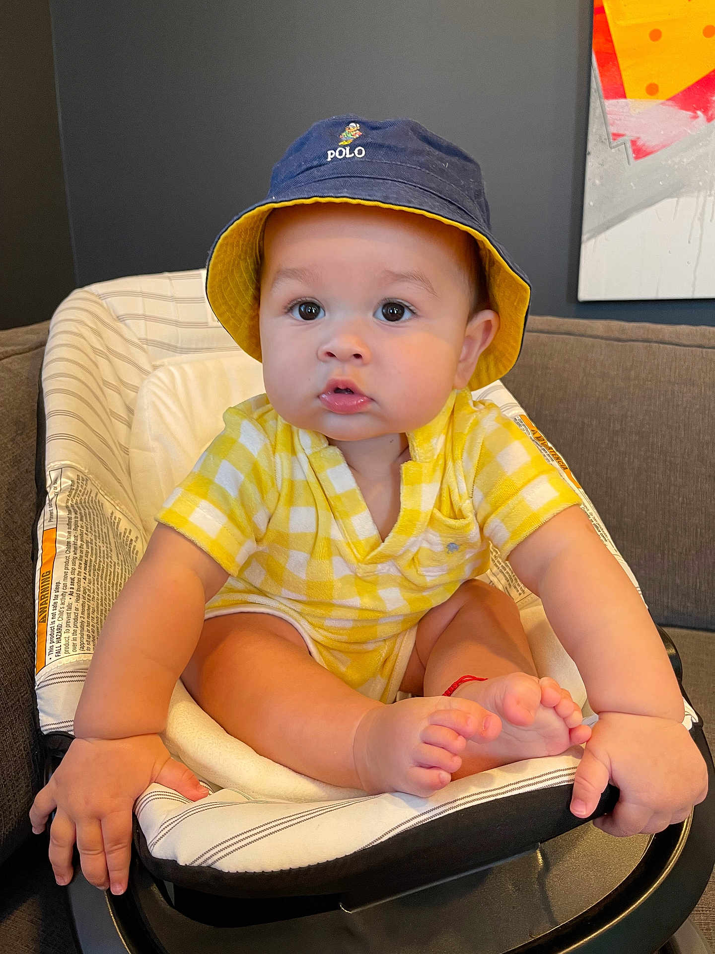 Carter joined the competition — help win amazing prizes! baby, child, hat, bucket_hat, yellow_clothing, baby_seat, indoor, cute, chubby_cheeks, hands, feet, polo_logo, plaid_pattern, seated, portrait, infant, couch, painting, home, curious