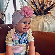 Ova a rejoint le concours — aidez-le/la à gagner de superbes lots ! baby, child, smiling, headband, flower, colorful_clothing, patchwork, couch, leather, indoor, vase, dried_plants, decor, window, curtains, happy, portrait, infant, sitting, cute