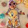 baby, child, tweety_bird, purple_outfit, flower_sunglasses, headwrap, tongue_out, blanket, paper_flowers, colorful, lying_down, cute, playful, smiling, indoors, soft_lighting, footwear, cartoon_character, flower_pattern, infant