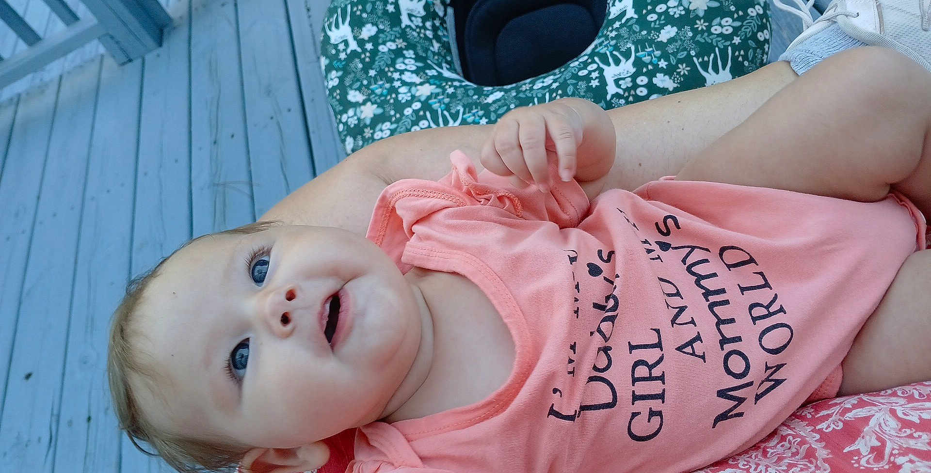 Emberly is registered to the contest to win money with this photo: baby, child, infant, smiling, blue_eyes, onesie, clothing, hand, face, skin, outdoor, deck, cushion, pattern, person, portrait, happy, young, cute, lying_down