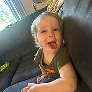 Luka is registered to the contest to win money with this photo: blanket, casual, child, clothing, couch, daylight, furniture, hair, happy, indoor, lamp, living_room, person, playful, sitting, skin, smiling, toddler, tongue_out, window