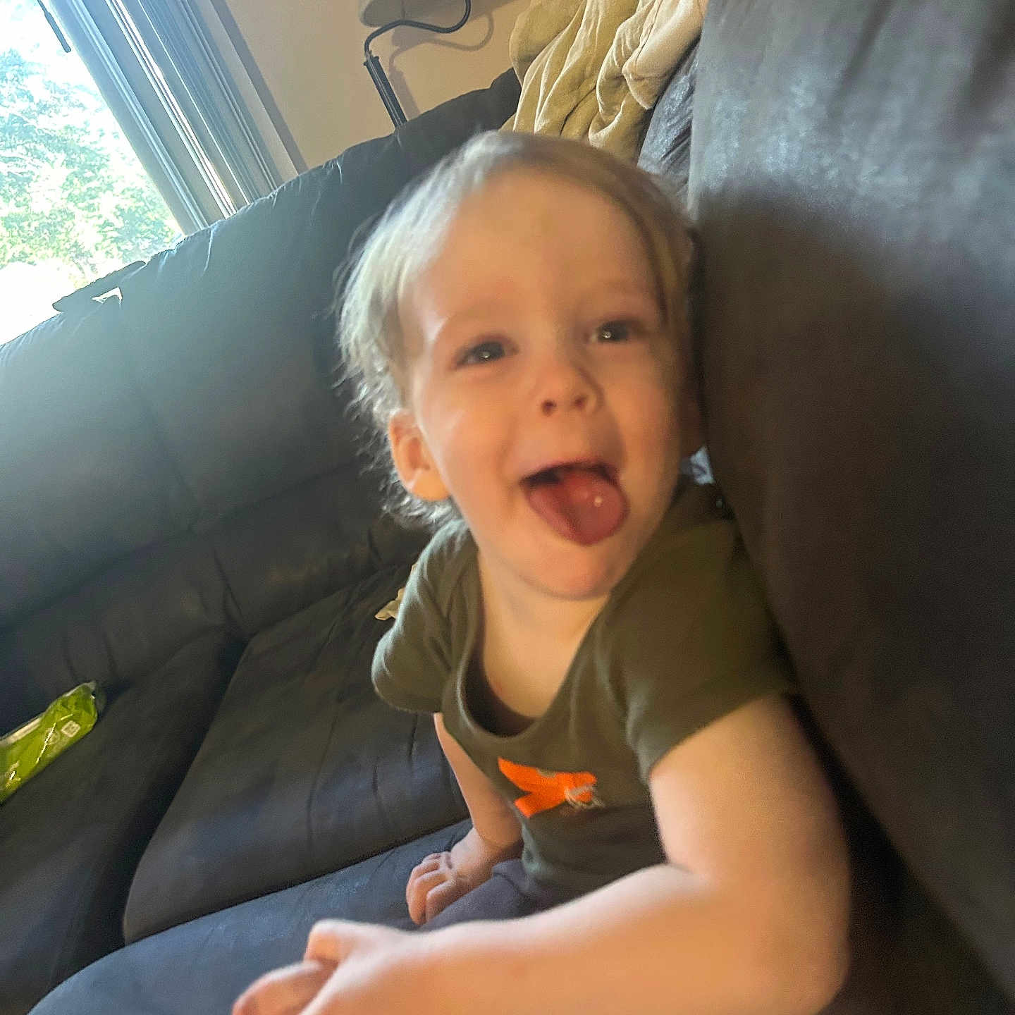 Luka is registered to the contest to win money with this photo: blanket, casual, child, clothing, couch, daylight, furniture, hair, happy, indoor, lamp, living_room, person, playful, sitting, skin, smiling, toddler, tongue_out, window