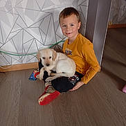 Aby a rejoint le concours — aidez-le/la à gagner de superbes lots ! boy, puppy, dog, child, indoor, floor, sitting, smiling, yellow_shirt, red_shoes, geometric_wall, cables, home, pet, young, casual, portrait, light, wooden_floor, comfort
