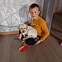 boy, puppy, dog, child, indoor, floor, sitting, smiling, yellow_shirt, red_shoes, geometric_wall, cables, home, pet, young, casual, portrait, light, wooden_floor, comfort