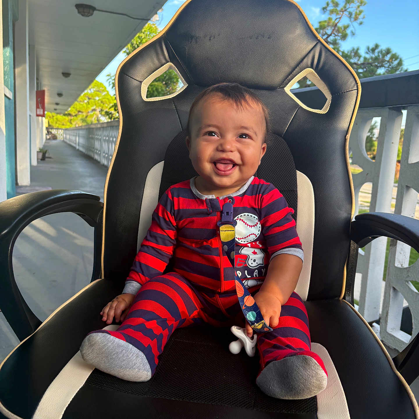 Giovanni is registered to the contest to win money with this photo: baby, child, smiling, pajamas, striped_clothing, pacifier, chair, gaming_chair, outdoor, porch, railing, daylight, happy, person, infant, footwear, sock, seat, expression, cute