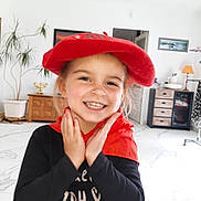 Léa participe au concours pour gagner de l'argent avec cette photo : child, smiling, red_beret, red_scarf, black_shirt, indoor, living_room, plant, furniture, cabinet, drawer, painting, door, floor, tile, happy, portrait, person, hands, face