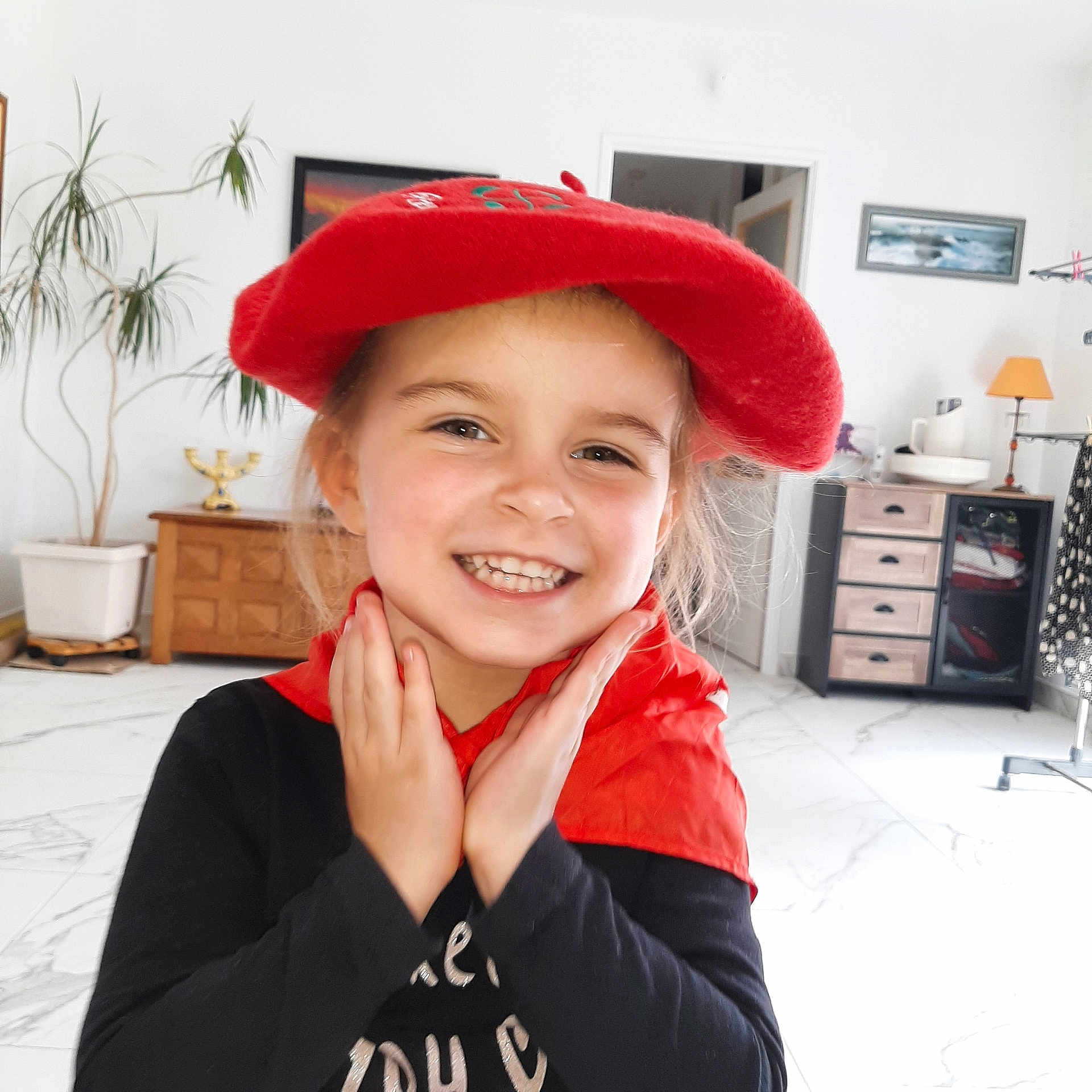 Léa participe au concours pour gagner de l'argent avec cette photo : child, smiling, red_beret, red_scarf, black_shirt, indoor, living_room, plant, furniture, cabinet, drawer, painting, door, floor, tile, happy, portrait, person, hands, face