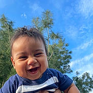 Matteo is registered to the contest to win money with this photo: baby, smiling, child, outdoor, sky, blue_sky, trees, greenery, happy, person, face, holding, toddler, short_hair, casual_clothing, blue_shirt, daylight, nature, portrait, cute