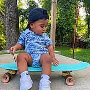 Matteo is registered to the contest to win money with this photo: toddler, child, skateboard, backwards_cap, white_sneakers, blue_outfit, outdoor, greenery, trees, concrete, grass, summer, young_child, casual_clothing, cap, playful, cute, side_profile, sunlight, nature