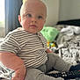 baby, child, indoor, bed, striped_shirt, gray_pants, remote_control, blue_eyes, sitting, cute, plush, soft_focus, patterned_bedspread, cozy, curious, person, clothing, furniture, room, portrait