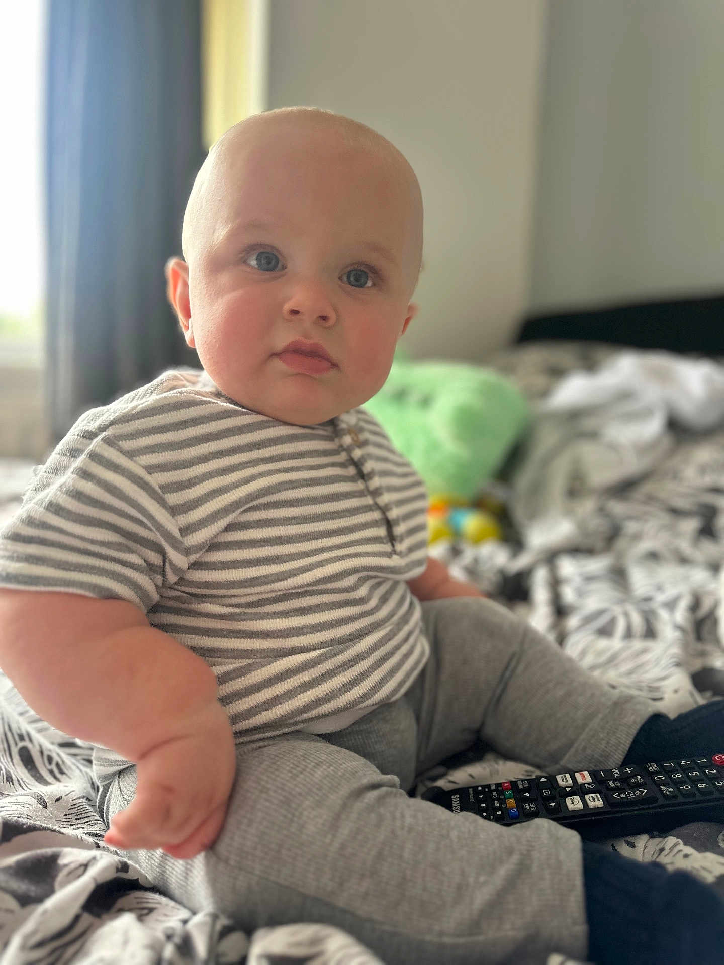 Cohen joined the competition — help win amazing prizes! baby, child, indoor, bed, striped_shirt, gray_pants, remote_control, blue_eyes, sitting, cute, plush, soft_focus, patterned_bedspread, cozy, curious, person, clothing, furniture, room, portrait