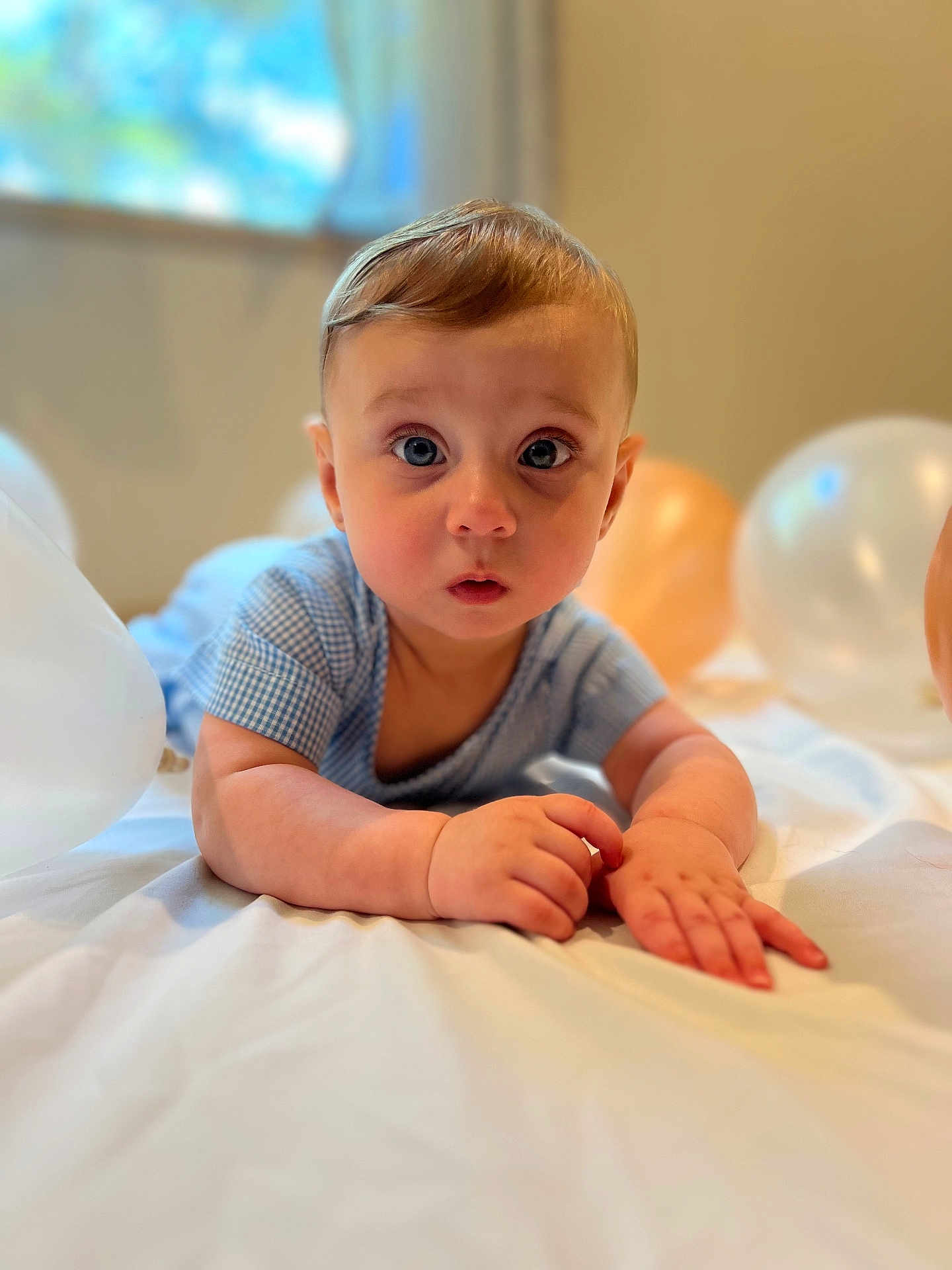 Levi is registered to the contest to win money with this photo: baby, child, infant, blue_outfit, balloons, curious_expression, lying_down, soft_surface, indoors, window, natural_light, cute, portrait, hands, face, young_child, short_hair, skin, head, eyes